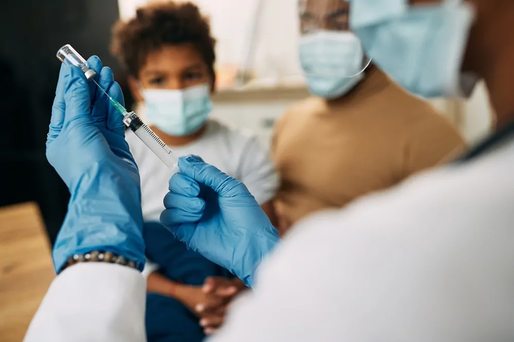 Em destaque mãos de mulher adulta com luvas azuis injetam a injeção em um vidrinho de vacina. Ao fundo criança com máscara assiste a preparação.