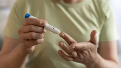 A imagem mostra um close-up das mãos de uma pessoa realizando um teste de glicemia capilar, procedimento comum para o monitoramento do diabetes.
