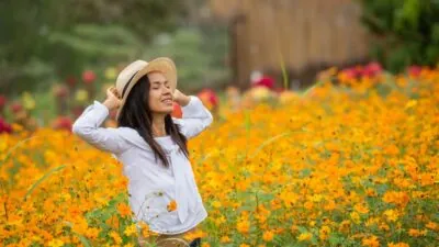 Mulher de chapéu sorri de olhos fechados em meio a campo de flores amarelas.