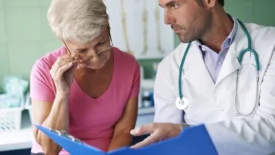 Doctor explains medical results to an elderly woman as they review a blue clipboard in a clinic setting, both focused on the information.